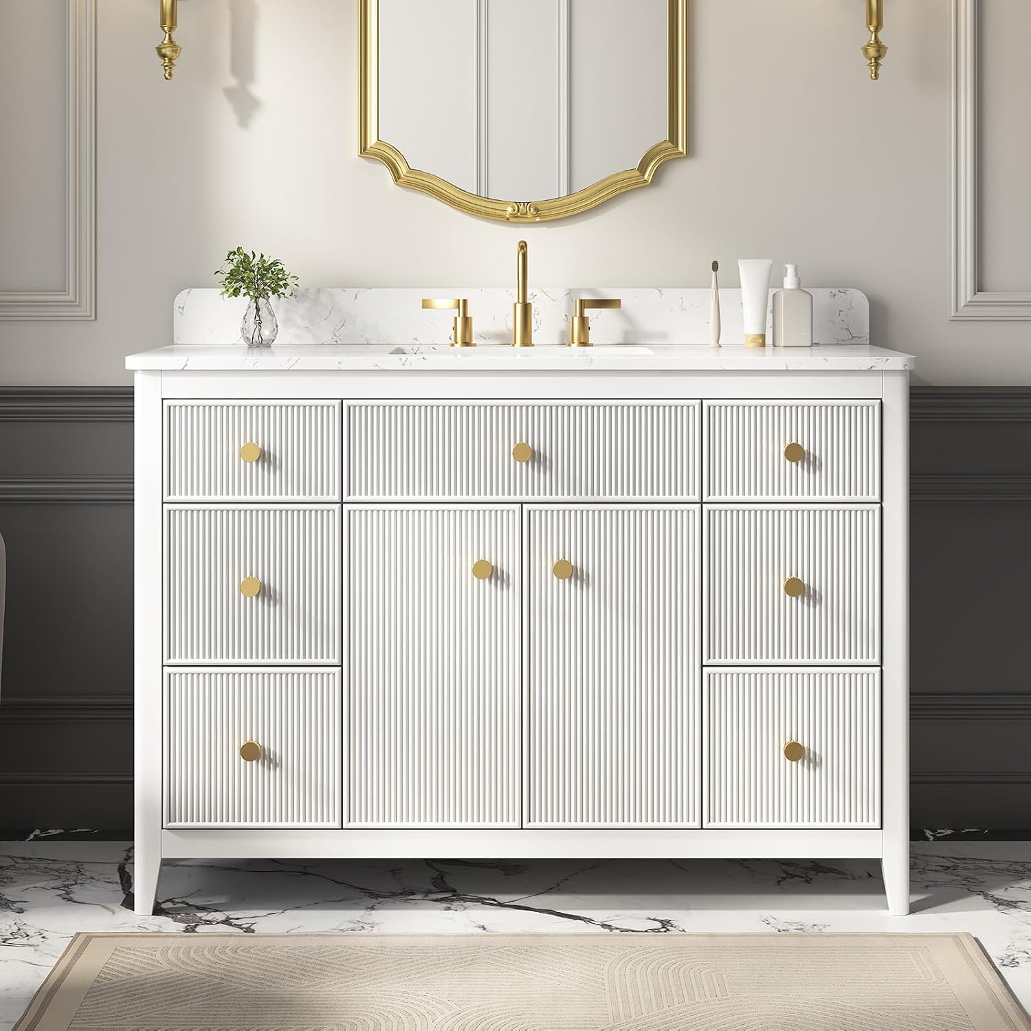A white bathroom vanity cabinet with vertical fluting, gold knobs, and a white marble top, shown in a styled bathroom setting with a gold mirror, gold faucet, and gray wainscoting.